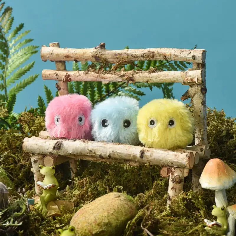 Three fluffy plush squeak toy balls for pets in pink, blue and yellow on a rustic log display with greenery.