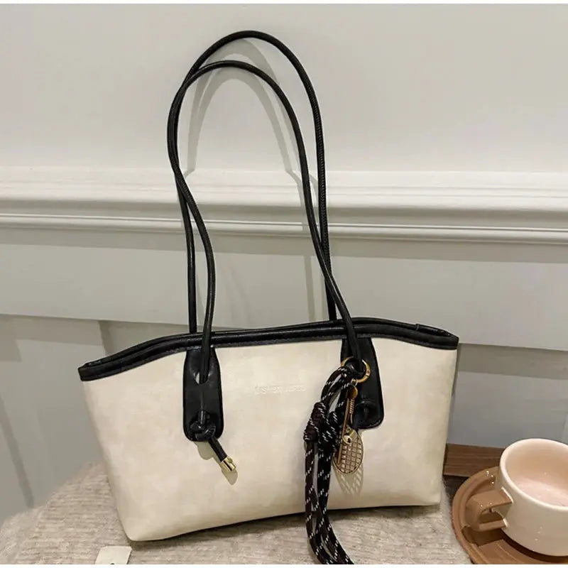 Beige canvas tote bag with black leather trim and long black handles, featuring a decorative pendant charm.