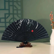 Black bamboo folding hand fan with white botanical pattern on wooden display stand