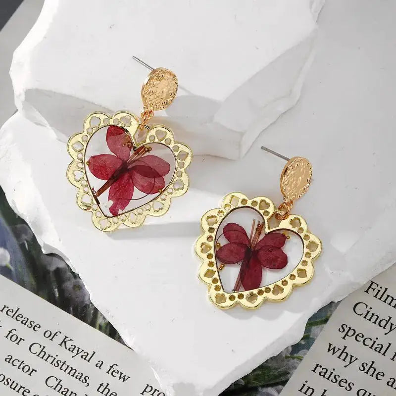 Pair of gold-tone heart-shaped dangle earrings featuring red pressed flowers inside clear epoxy resin.