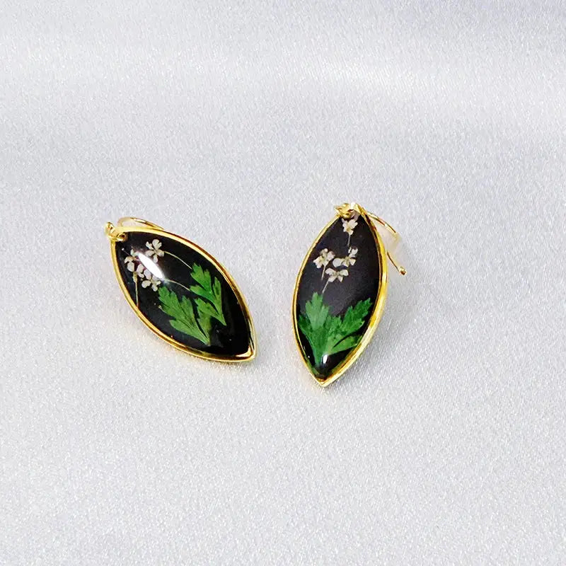 Pair of gold-edged teardrop epoxy resin earrings with pressed green leaves and white flowers on black background