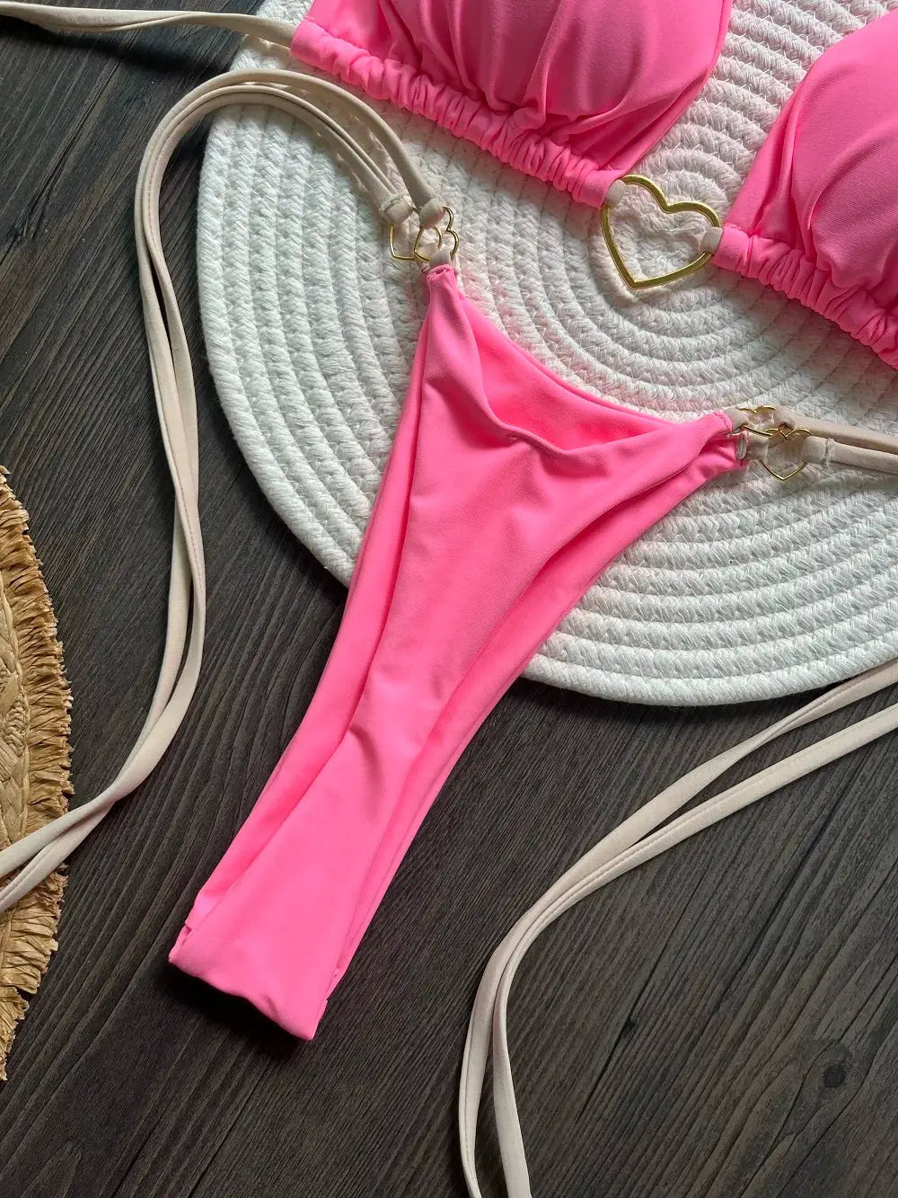 Pink two-piece halter bikini set with triangle top and thong bottom, beige straps with gold heart accents, on white mat.