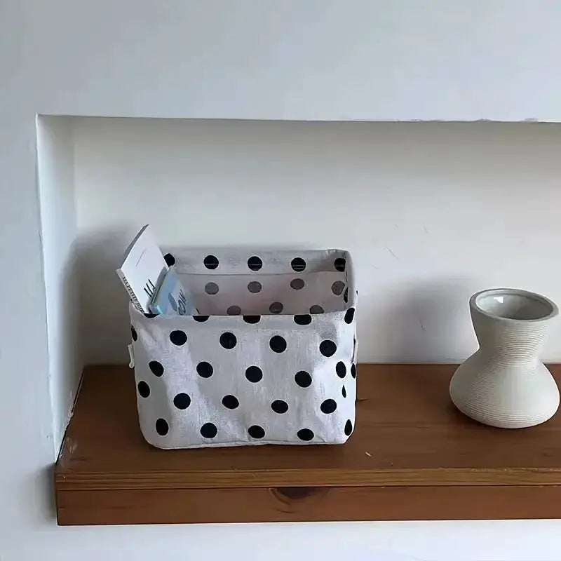 Small foldable canvas storage basket with handle, white fabric with black polka dots on a wooden shelf.