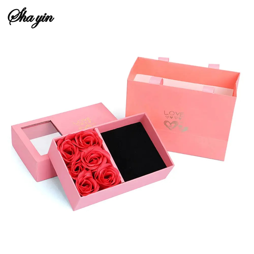 Pink jewelry box with red preserved roses and a black velvet insert; includes a matching pink gift bag.