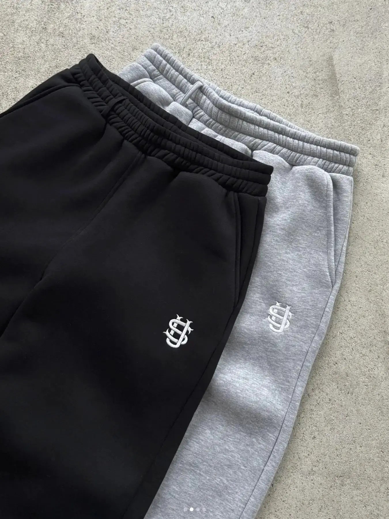 Two Simples Market joggers in black and gray with elastic waistbands and white embroidered logo on the thigh.