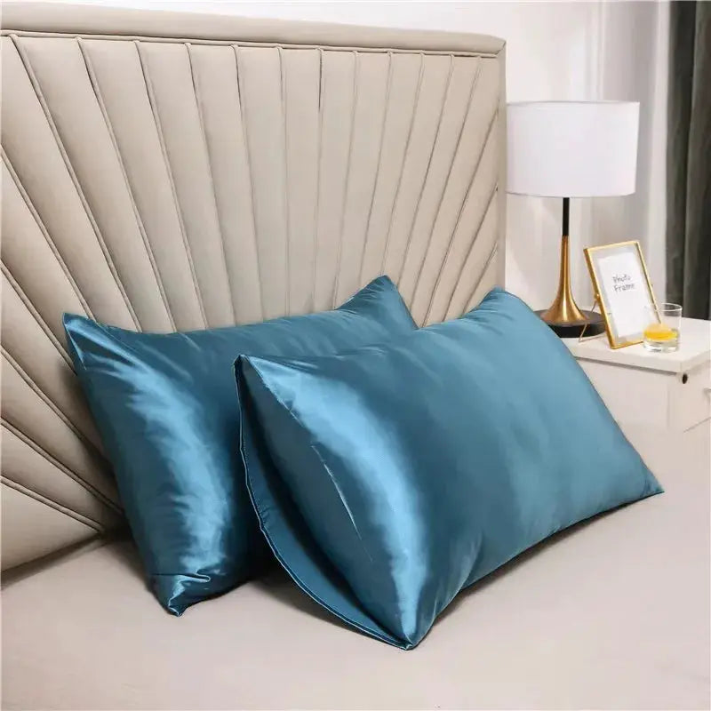 Two teal silk satin pillowcases on a beige upholstered headboard.
