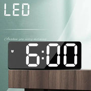 Digital LED alarm clock with a black mirrored panel displaying 6:00 on a wood bedside table