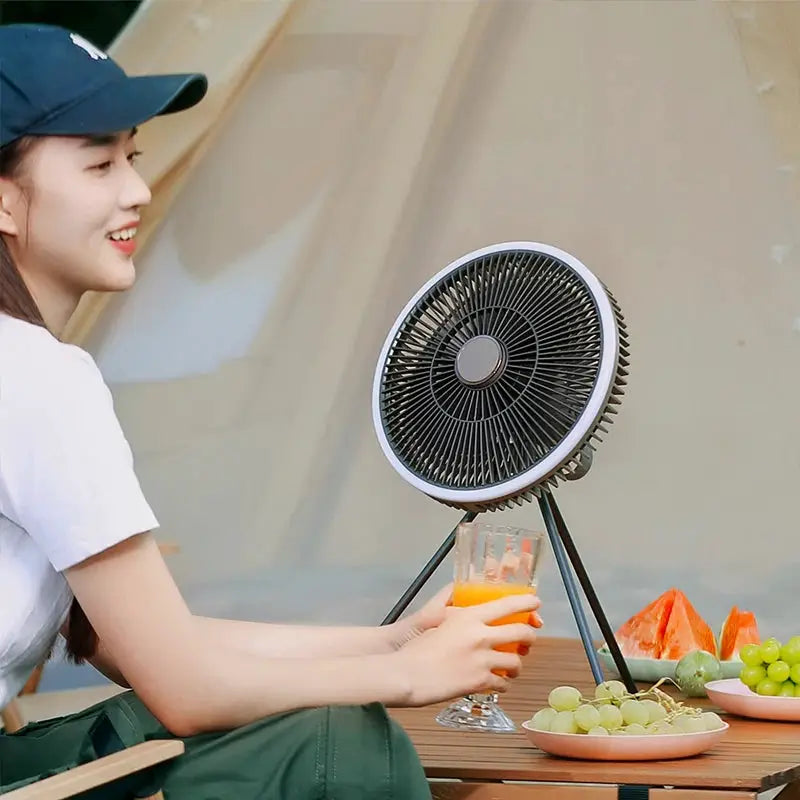 KINSCOTER outdoor camping fan on a tripod, pictured with a glass of orange juice and fruit.
