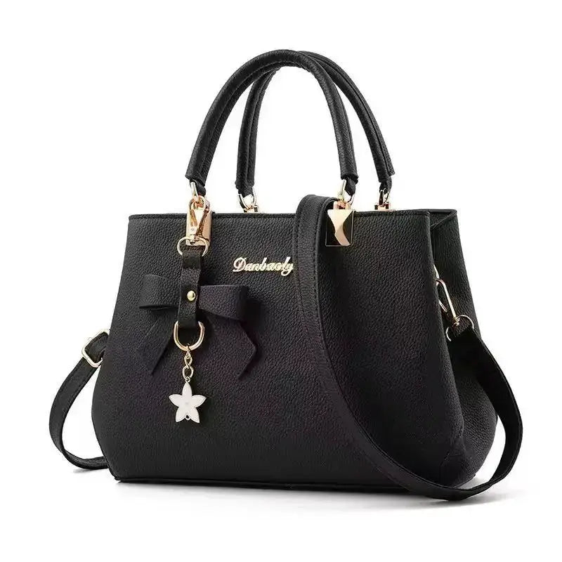 Black textured handbag by Simples Market with bow charm, gold logo plaque, double top handles, shoulder strap