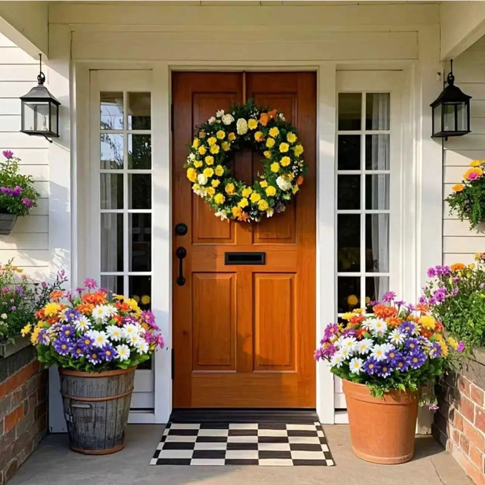 Artificial silk daisy wreath with yellow and white blooms on a front door, flanked by colorful potted flowers.