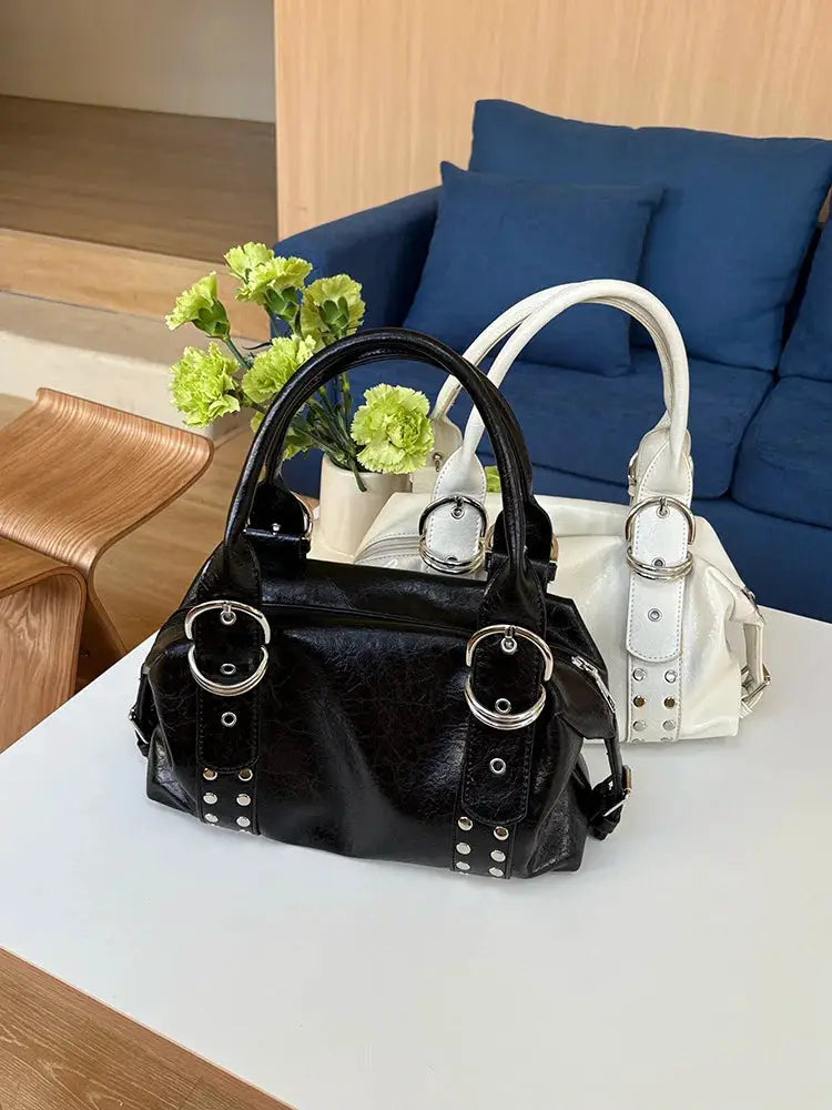 Two PU leather rivet shoulder bags in black and white with bold metal buckles on a white table.