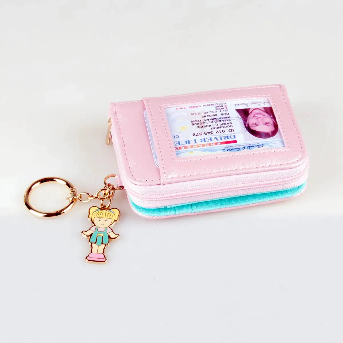 Pink stitched zipper wallet with ID window and turquoise accent, plus a gold keychain charm of a cartoon girl.