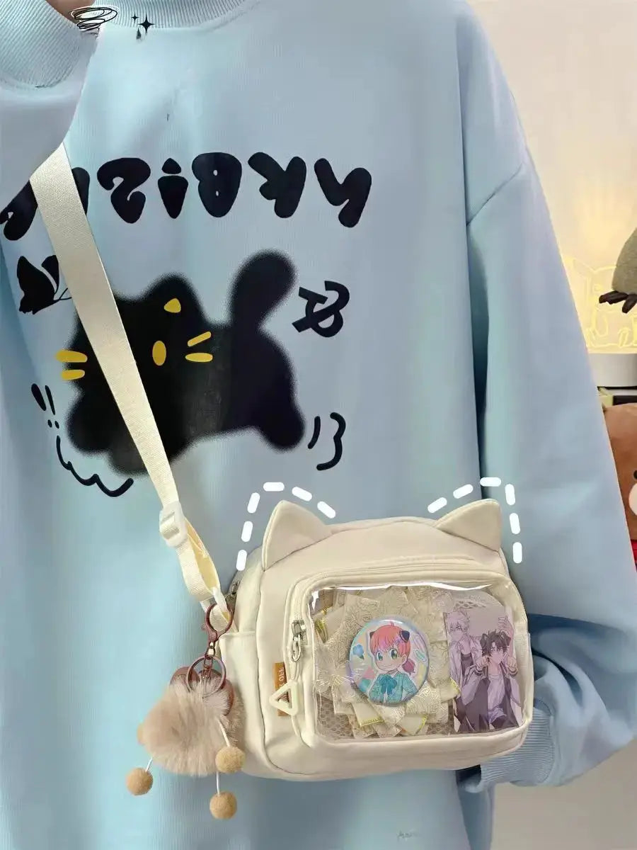 White cream Ita bag with cat-ear top, clear front pocket showing badges, crossbody strap and fluffy pom-pom keychain.