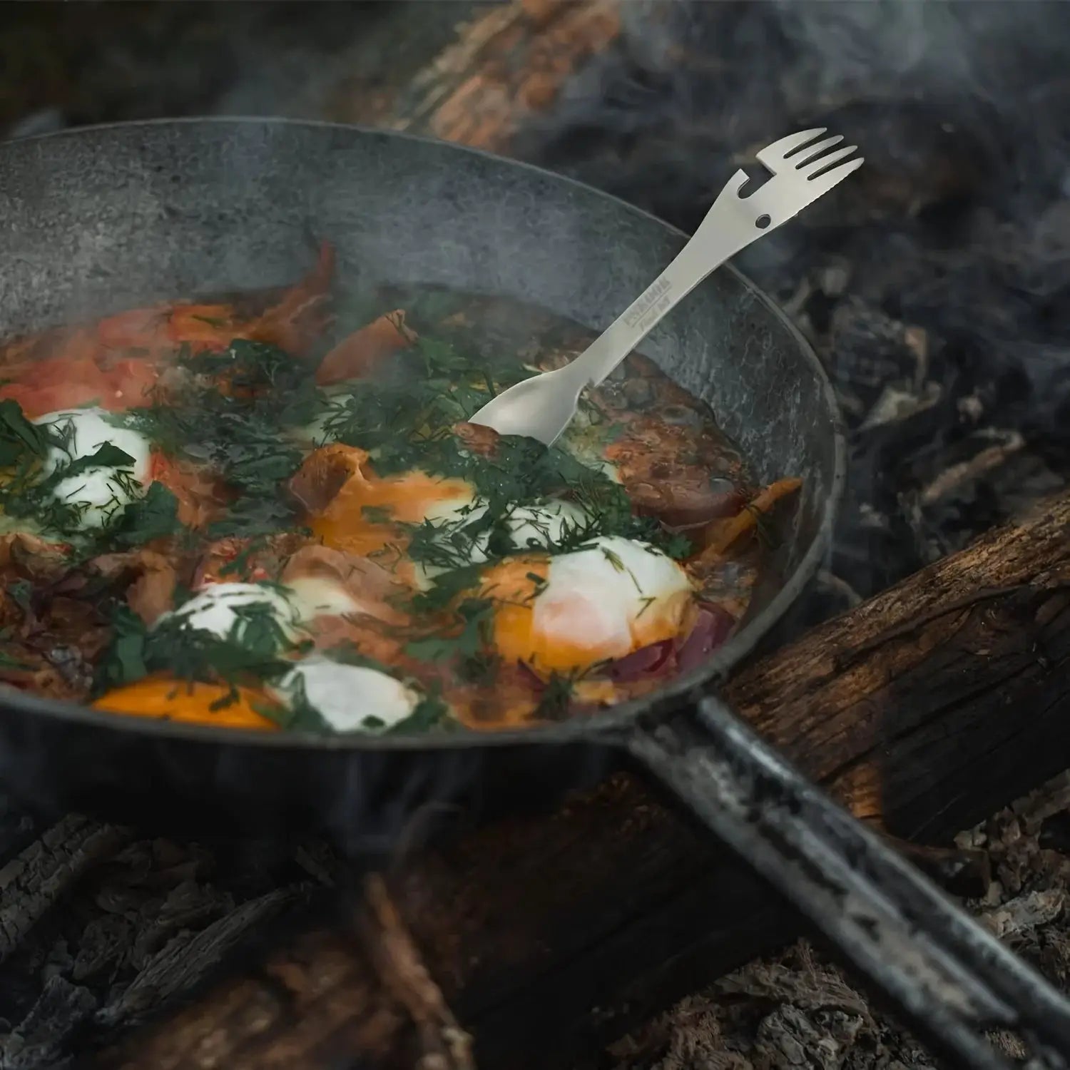 PSKOOK 5-in-1 camping multi-tool in stainless steel, resting in a skillet with eggs and herbs over a campfire