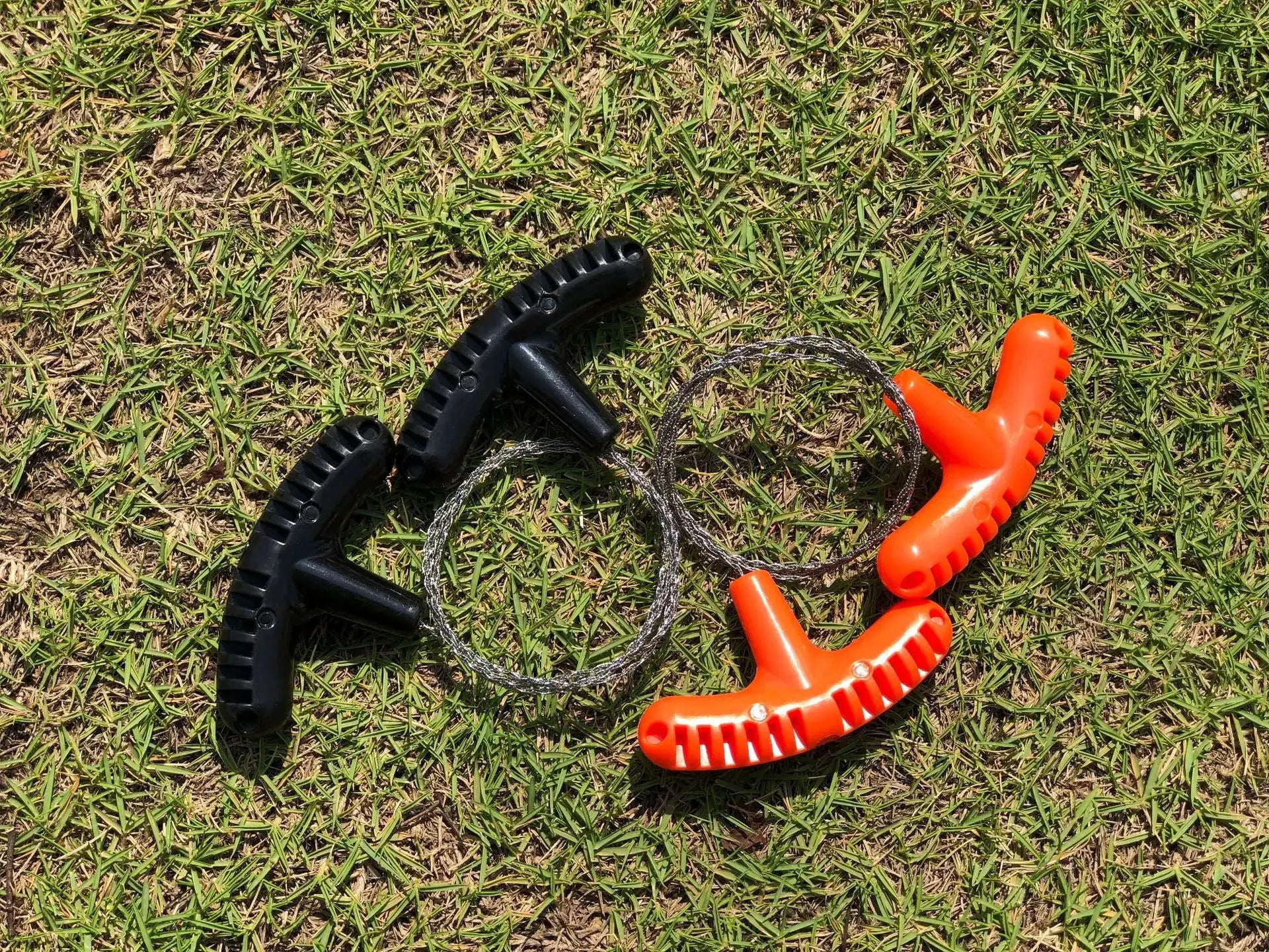 Manual rope chain saw with black and orange handles and braided steel wire on grass