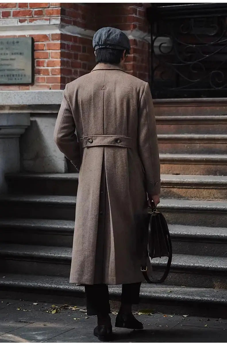 Beige wool-blend long men's overcoat with waist belt and back vent, carried with dark leather briefcase.