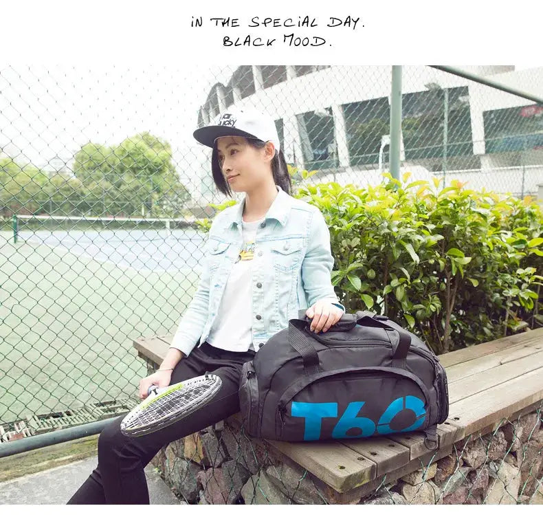 Gym bag by Simples Market, black duffel with blue T60 logo, outdoors beside a tennis court.