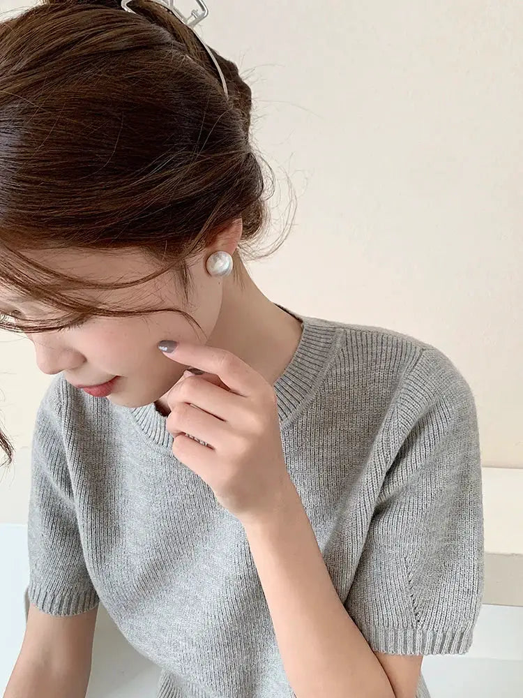 Pearl stud earrings with large white pearls on silver-tone posts, worn by a woman in a gray knit top.