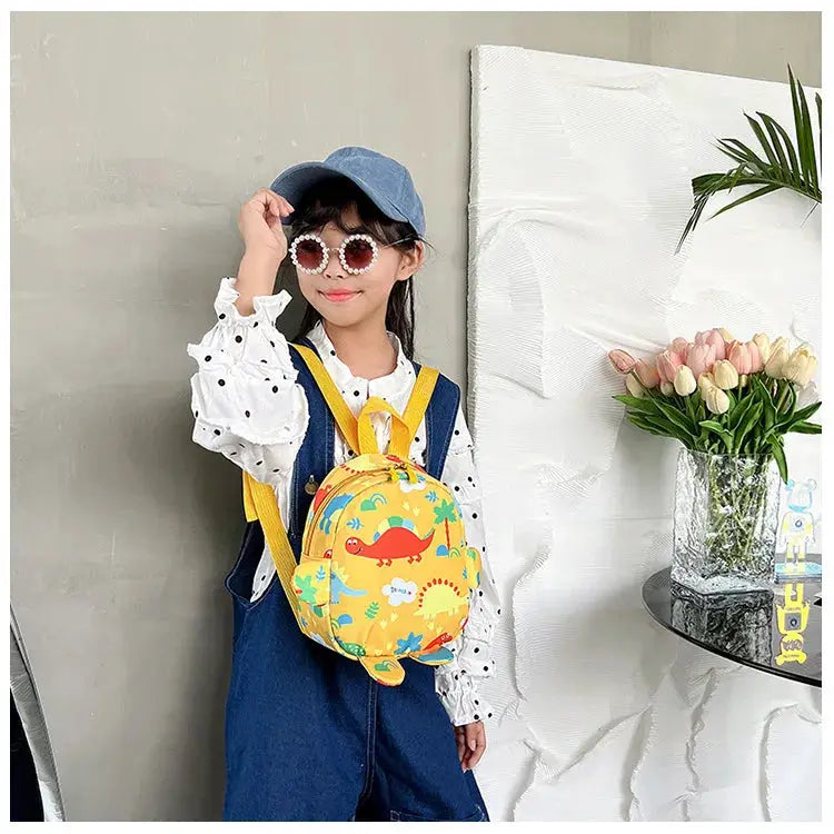 Kids cartoon dinosaur backpack, yellow with colorful dinosaur prints, worn by a girl in denim overalls.