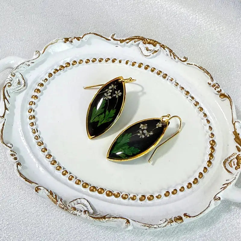 Pair of marquise-shaped epoxy resin earrings with pressed white flowers and green leaves, gold trim on black background