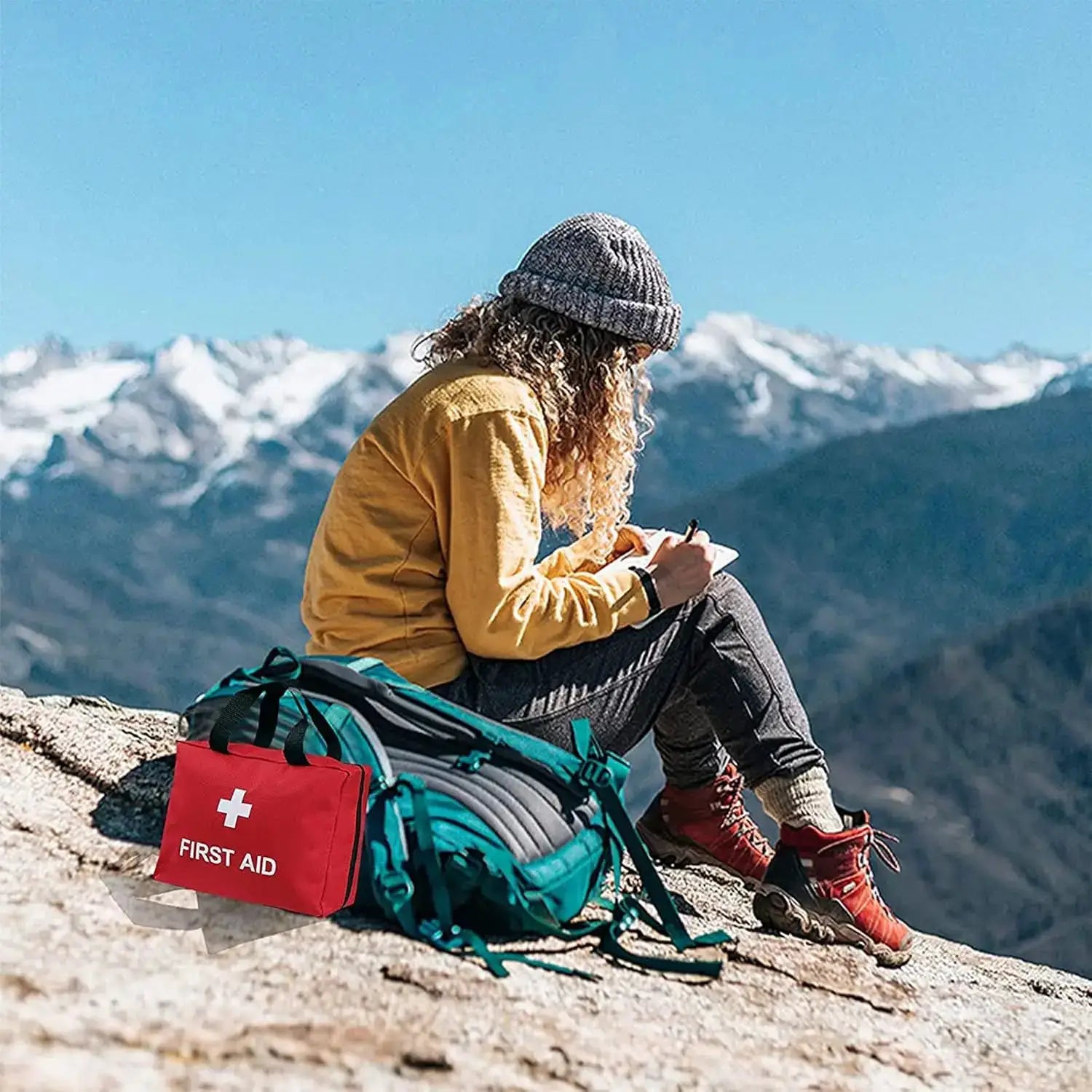 260pcs Deluxe First Aid Kit in red bag with white cross, on rocky mountain trail beside a teal backpack.
