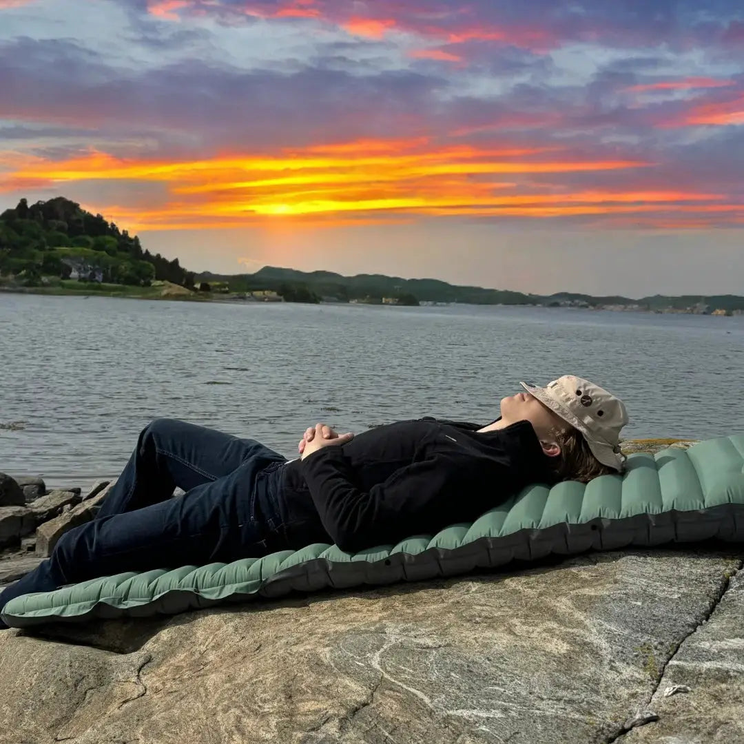 Simples Market extra-thick 4-inch camping sleeping pad in green, self-inflating, laid on rocks by a lake at sunset