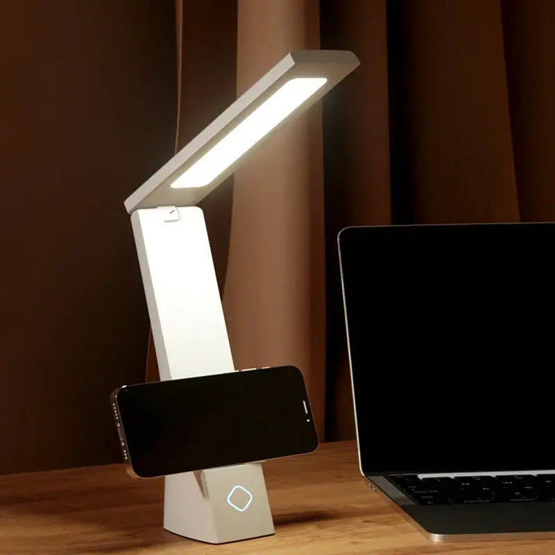 White LED desk lamp with folding arm and phone charging stand for eye protection beside laptop
