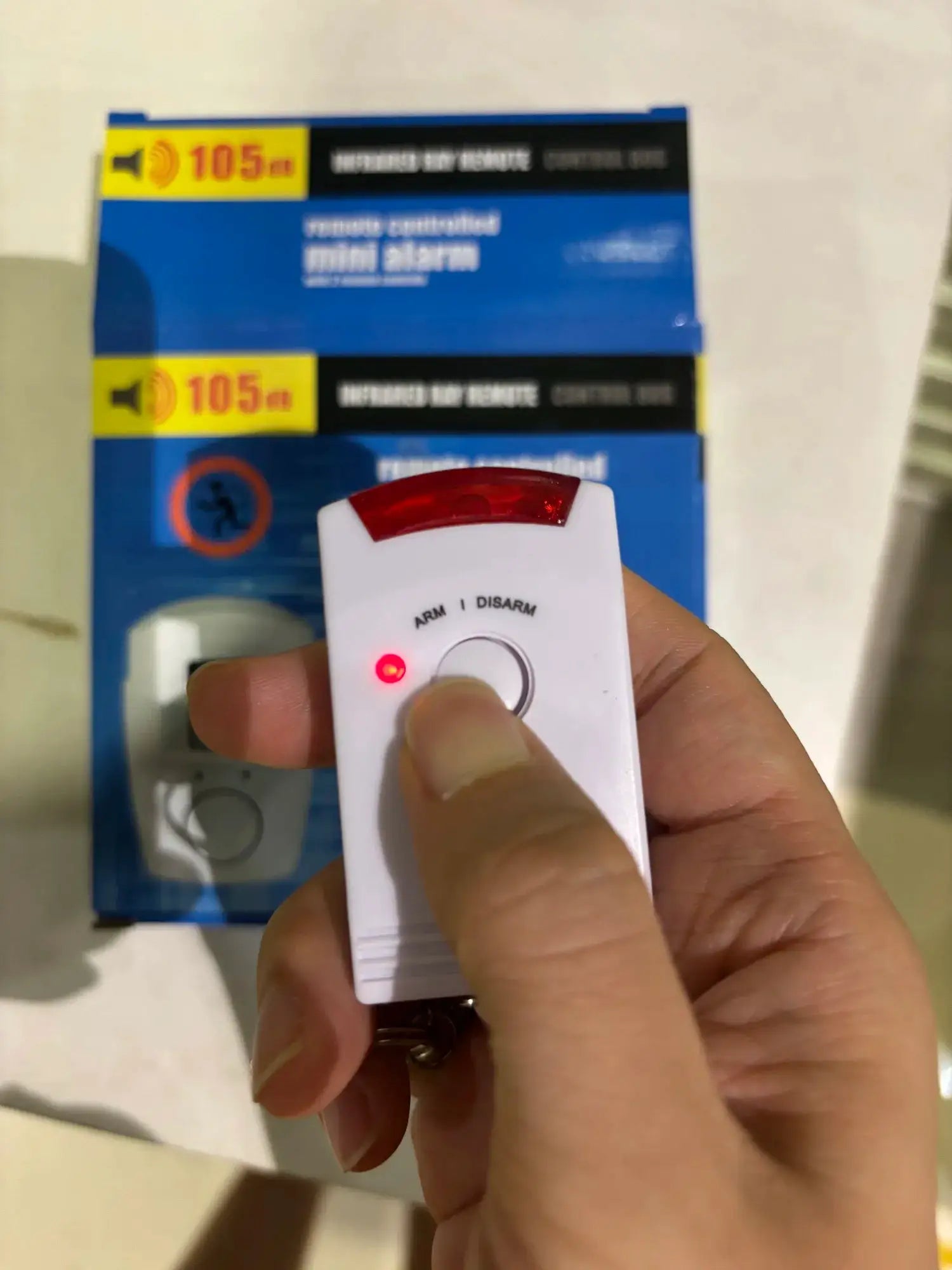 Wireless motion sensor alarm remote held in hand; white device with red LED in front of blue 105 dB packaging.