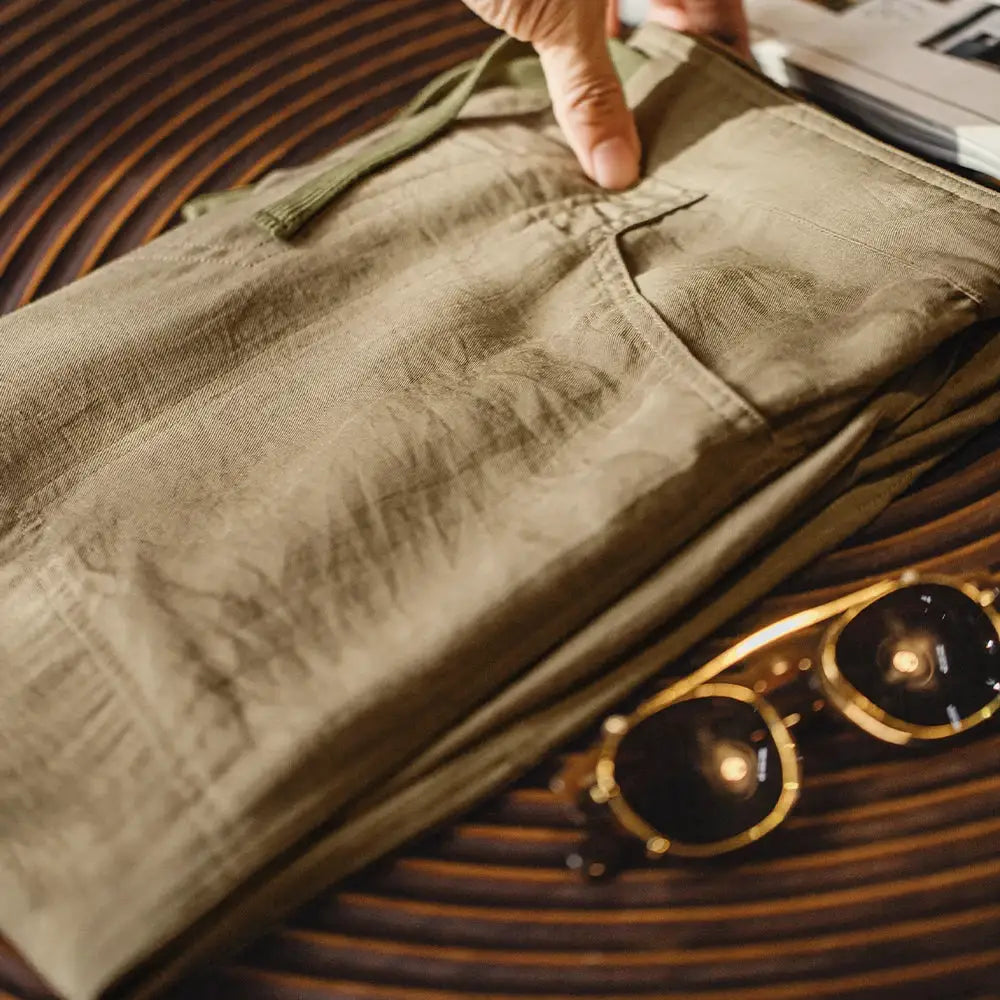 Maden men's linen-blend chino pants in khaki with elastic waist, folded on wood beside sunglasses