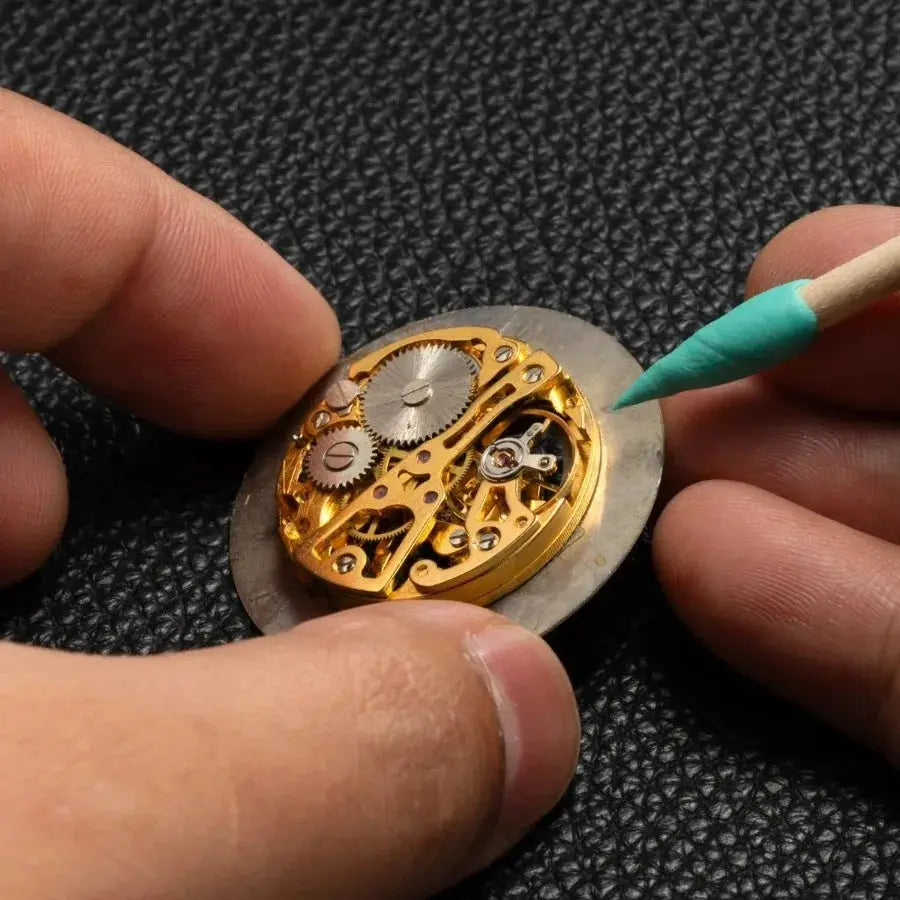 Watch cleaning tool: gold-toned mechanical movement held by fingers with a green-tipped precision cleaner