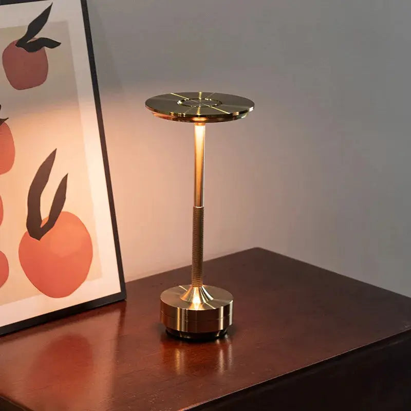 Gold-tone wireless decorative table lamp with round top, infinitely dimmable by touch, on a dark wood surface.