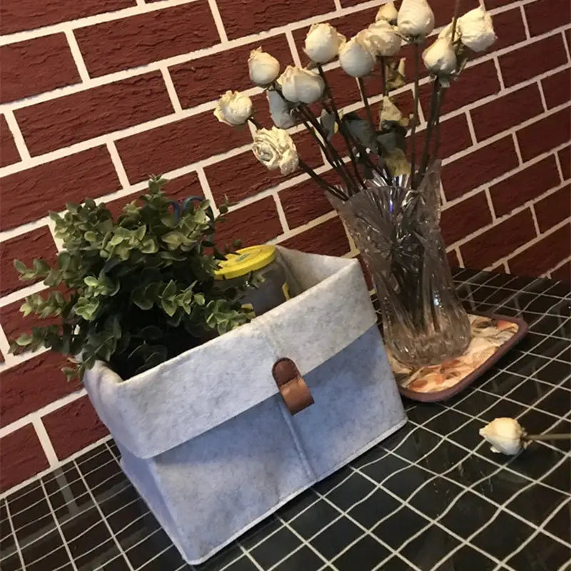 Blue-gray felt storage basket and glass vase with white roses on a grid surface.