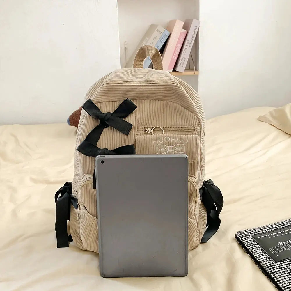 Beige corduroy backpack with black bow accents and a front zipper pocket, tablet for scale.