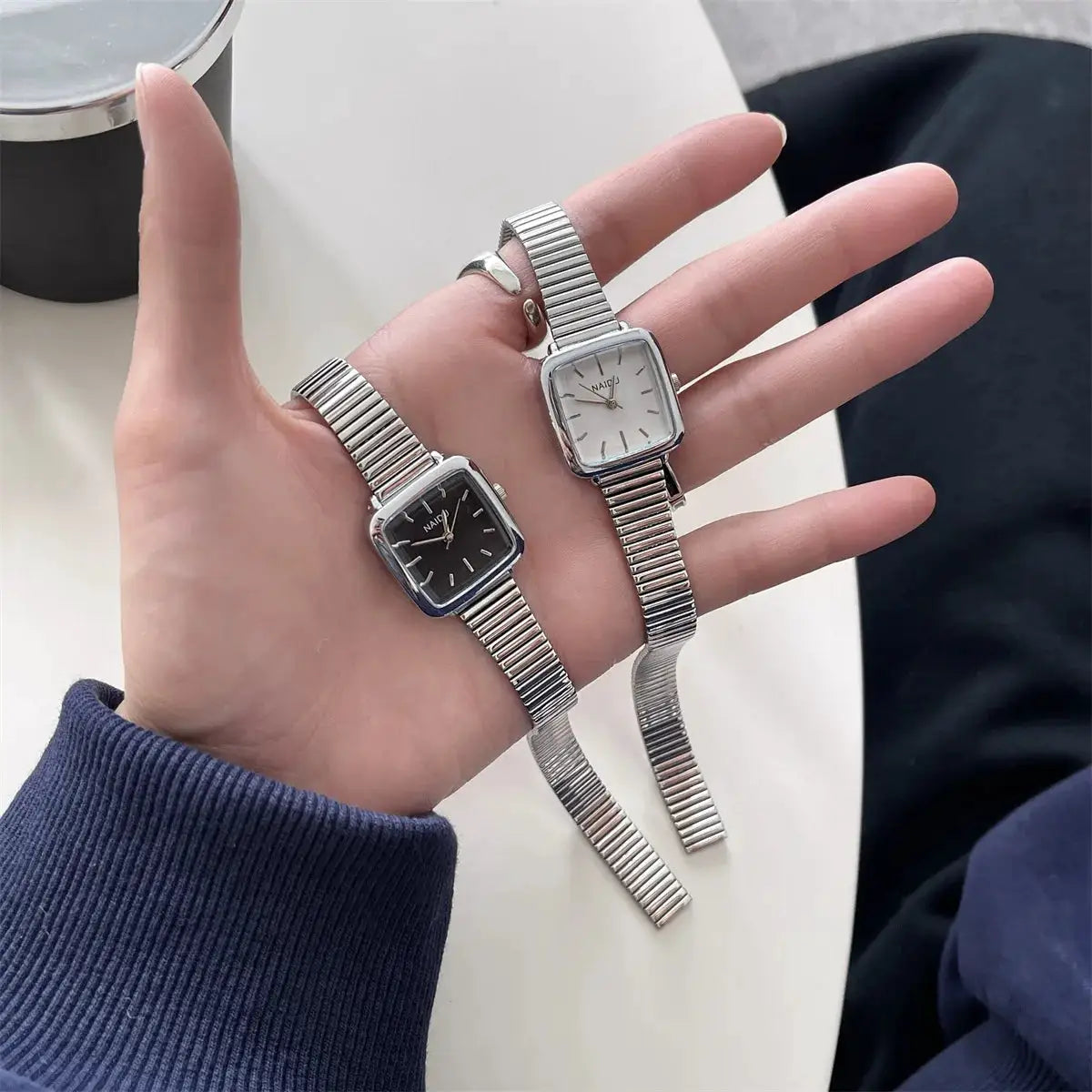 Two women's square-face quartz watches with stainless steel stretch bands; one black dial, one white dial.