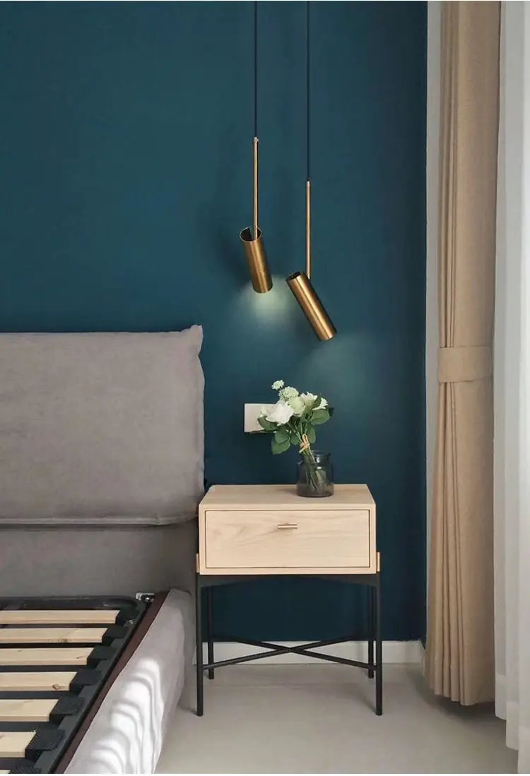 Minimalist LED pendant lights, two brass cylindrical pendants with warm glow over teal wall beside nightstand