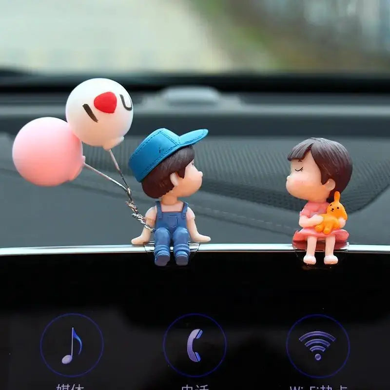 Car dashboard ornament: two cartoon couple figurines, blue-overall boy with balloons and pink-dress girl with cat