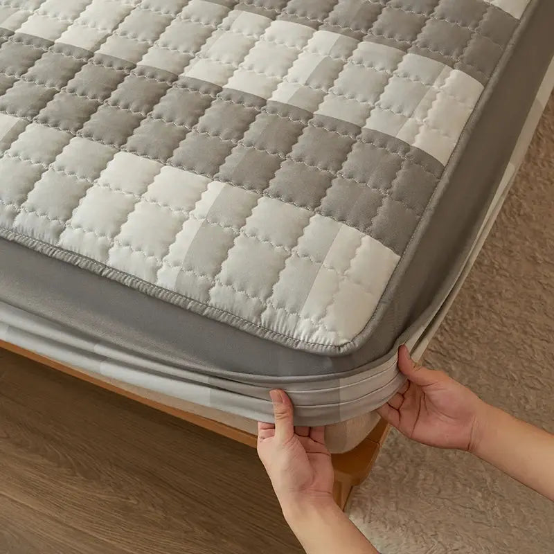 Quilted plaid mattress protector in grey and white being fitted to a bed, with a soft grey border.