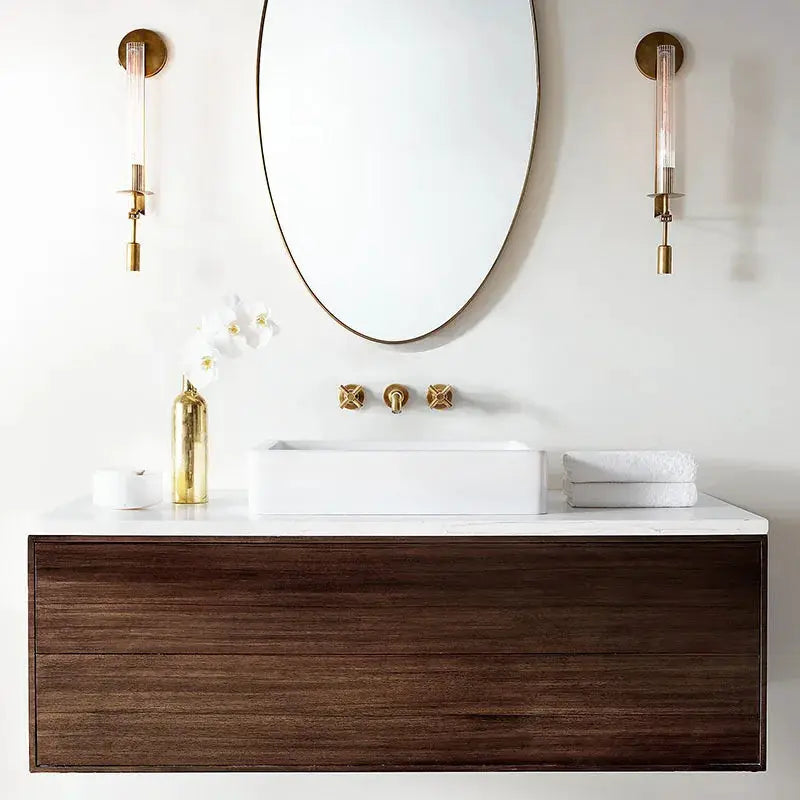 Modern copper wall sconces (2) with clear glass tubes beside a round mirror over a white vessel sink on a wood vanity