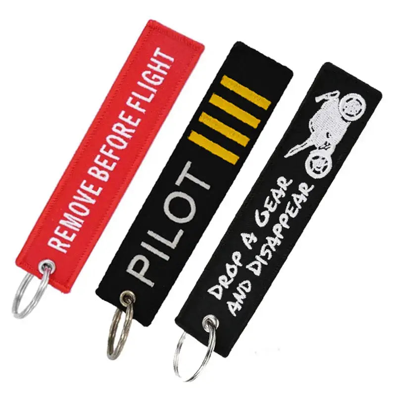 Three embroidered fabric key fobs with metal rings: red REMOVE BEFORE FLIGHT, black PILOT, and black with a cyclist graphic.