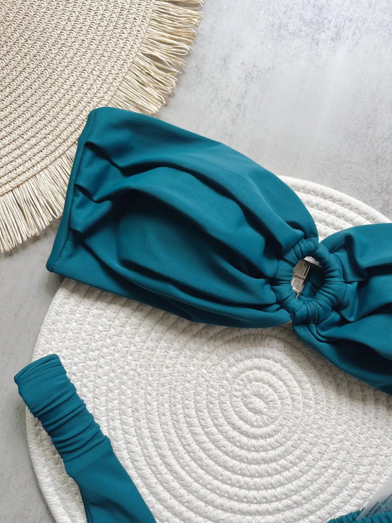 Teal micro bikini top with ruched center and metal hardware, laid on a white woven mat.