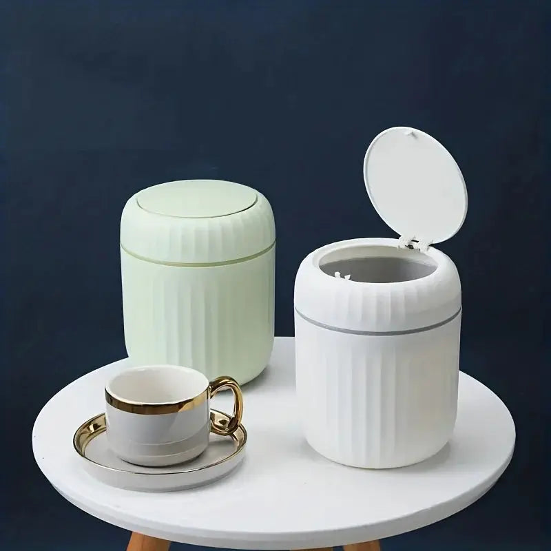 Two mini desktop trash cans with lids, ribbed white and pale green plastic, on a round table beside a teacup.