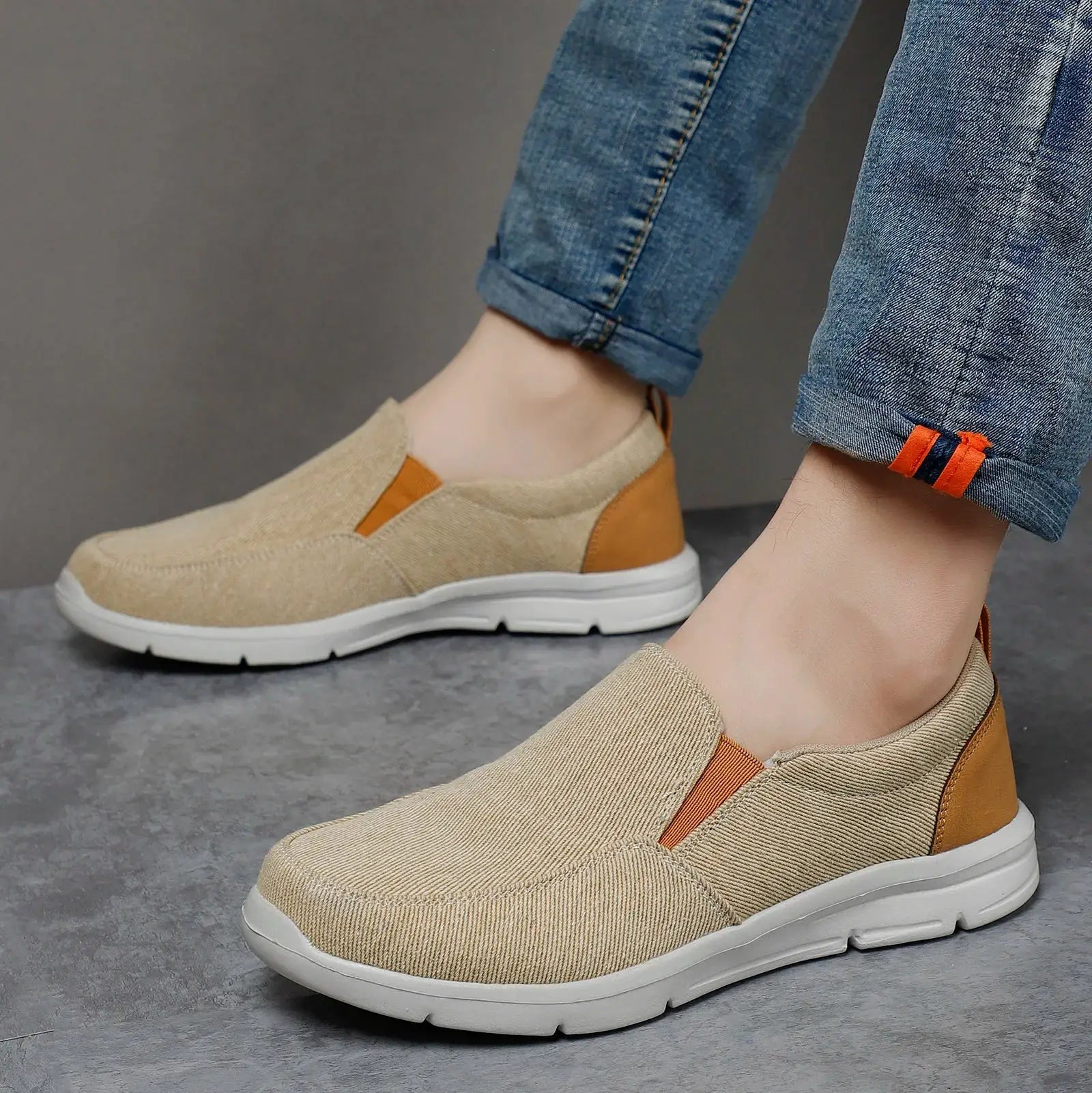 Pair of men's beige canvas slip-on sneakers with brown heel panel and orange pull tab, white sole