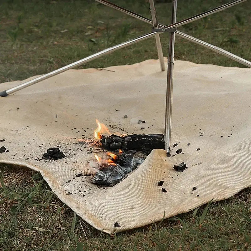 Fireproof camping insulation mat in beige on grass under a metal camping umbrella with a small campfire.