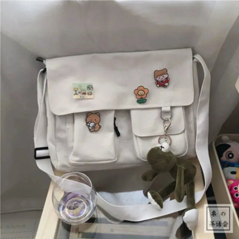 White canvas messenger bag with flap, front pockets and cartoon patches; crossbody strap.