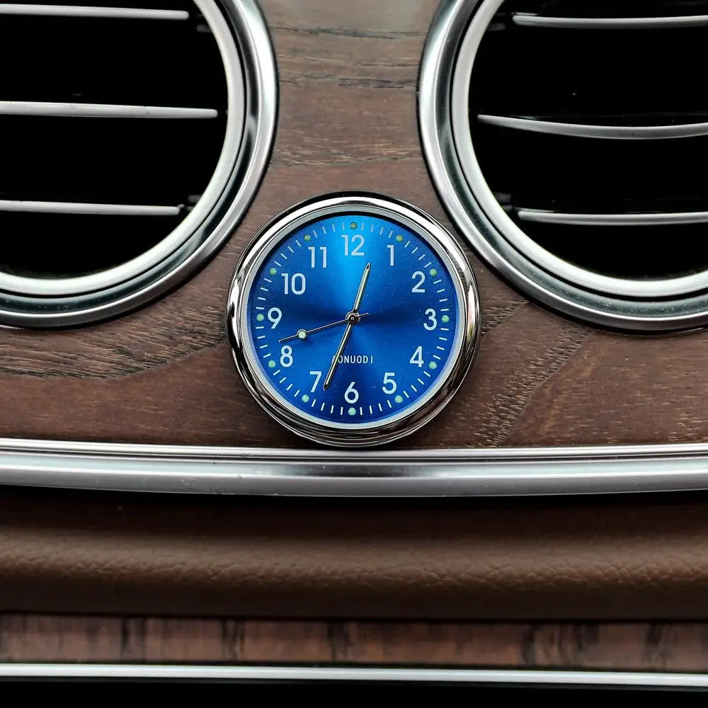 Car clock with luminous blue dial and chrome bezel, mounted on a woodgrain dashboard between two round air vents