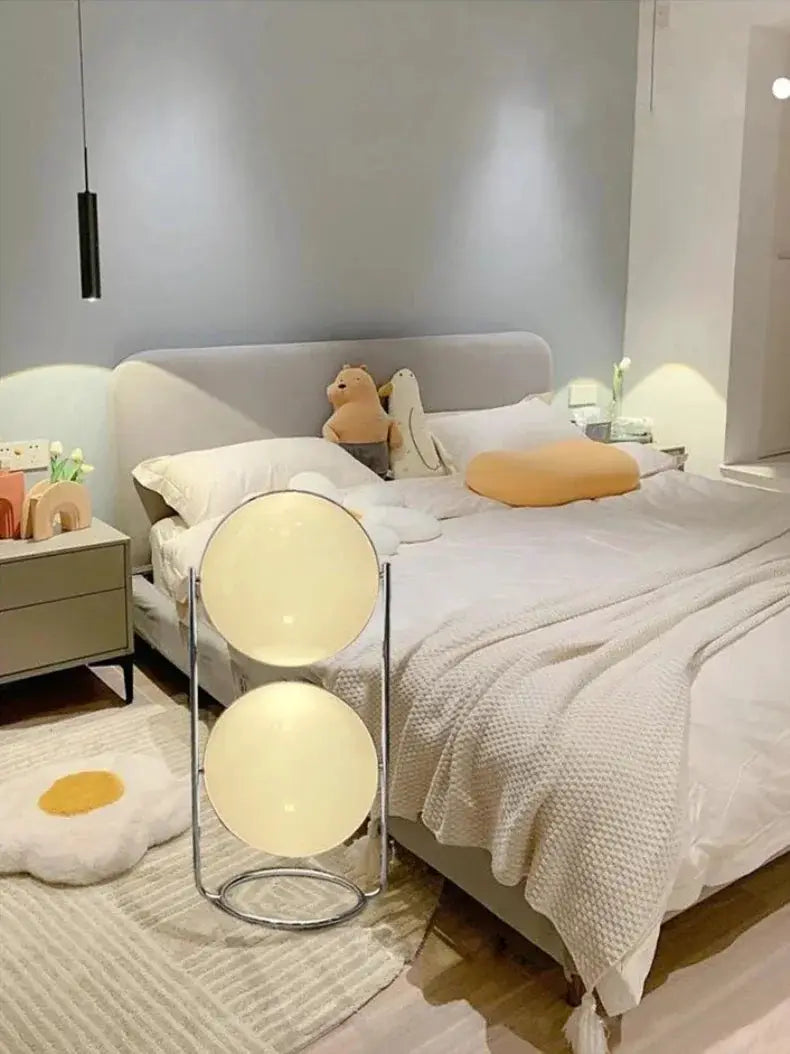 Modern white acrylic circular LED floor lamp with double globe shades beside bed.