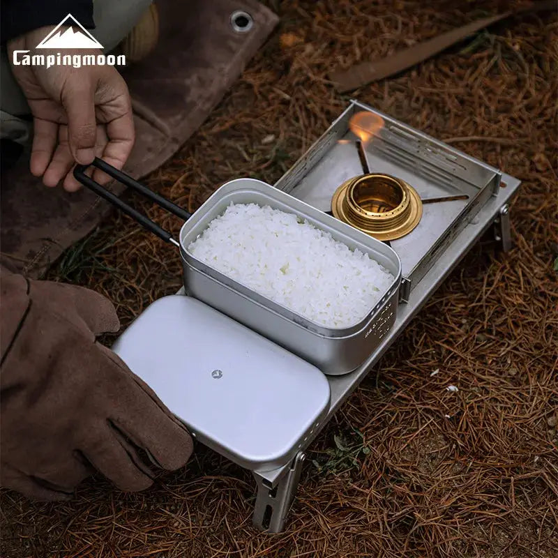 Campingmoon aluminum lunch box on a portable stove outdoors, rice-filled with lid nearby.