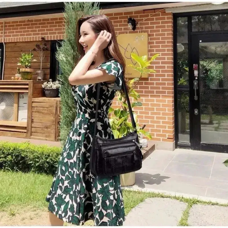 Crossbody shoulder bag in black with multiple zip pockets, worn by a woman in a green leaf-print dress.