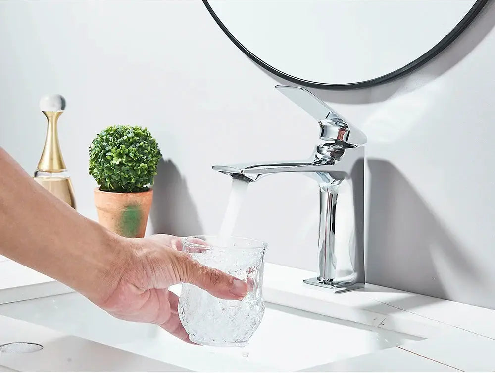 Deck-mounted chrome bathroom sink mixer tap with a single lever; water flows into a glass.
