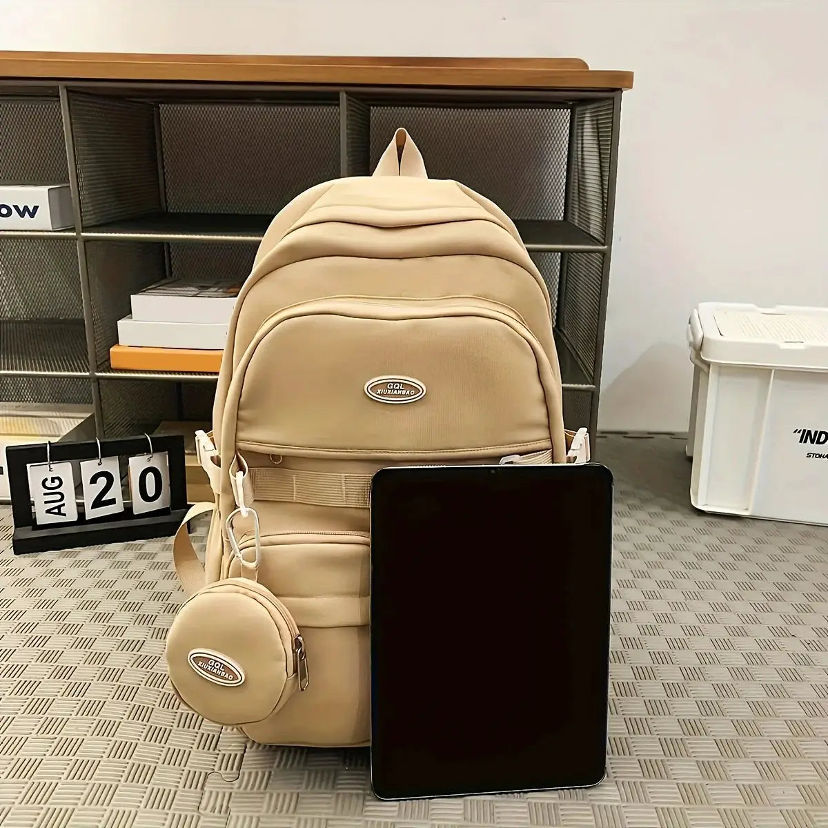 Beige large-capacity travel backpack with front pocket and attached round mini pouch, shown with tablet.